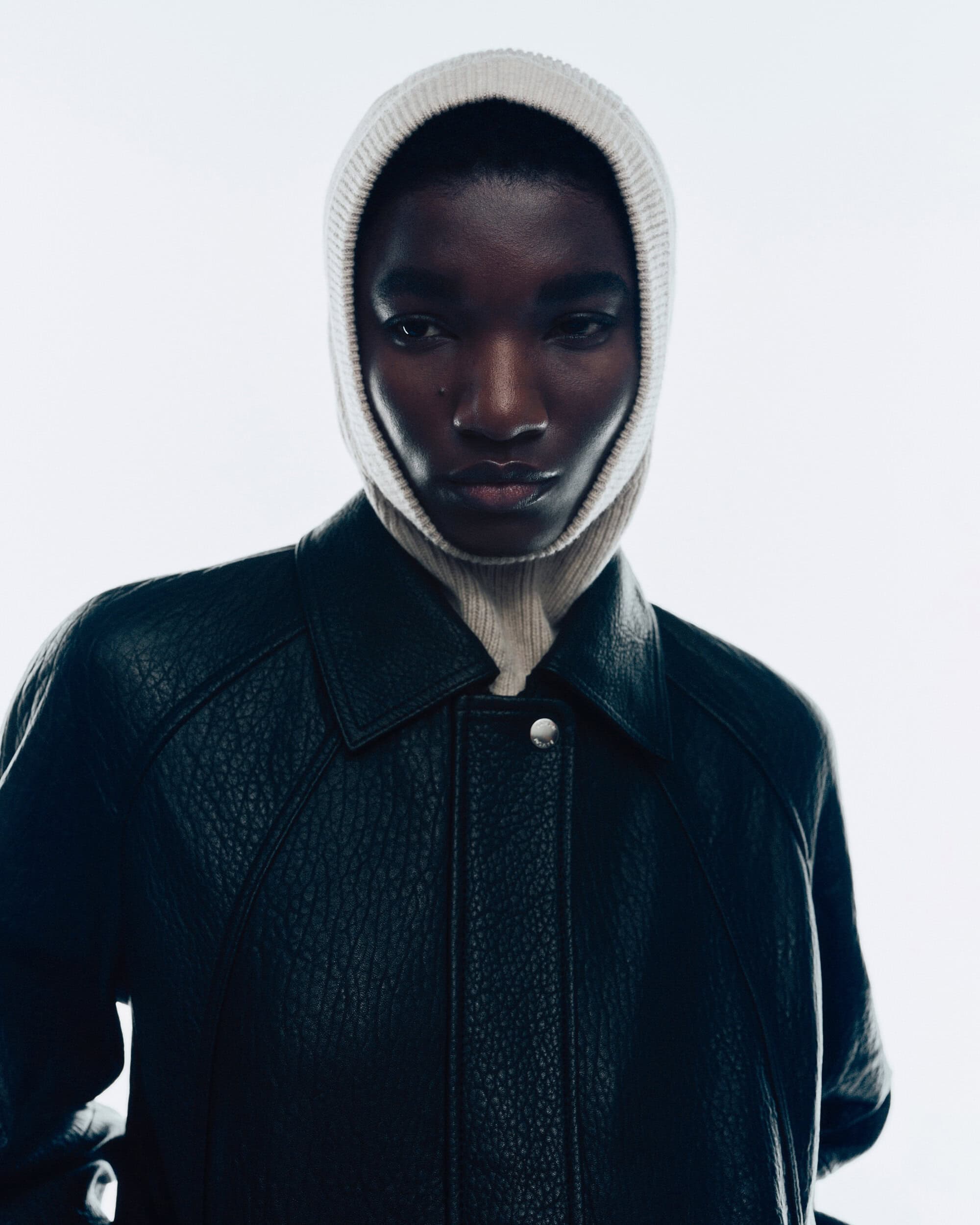 Model wearing a white ribbed knit balaclava and dark leather jacket against a bright white background for Samsoe Samsoe campaign