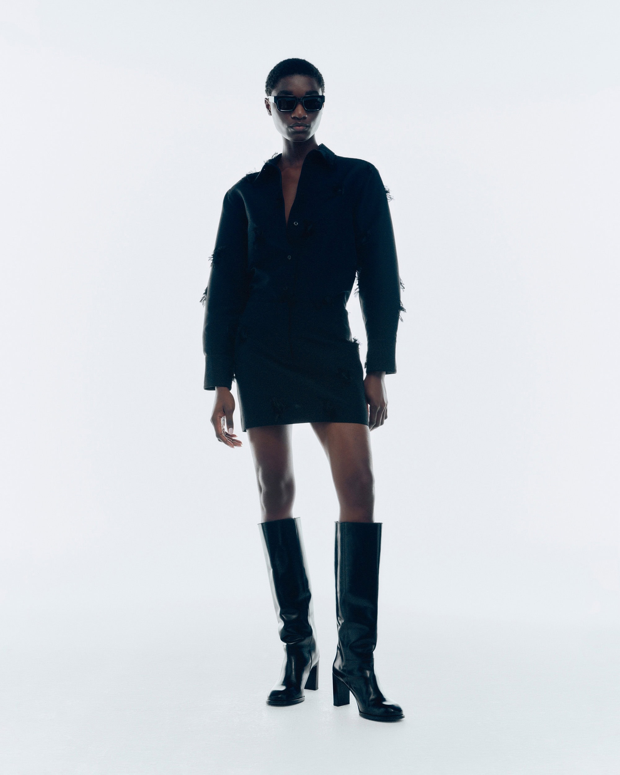 Model in black sunglasses, dark button-down shirt, matching skirt, and tall boots against a white studio background.