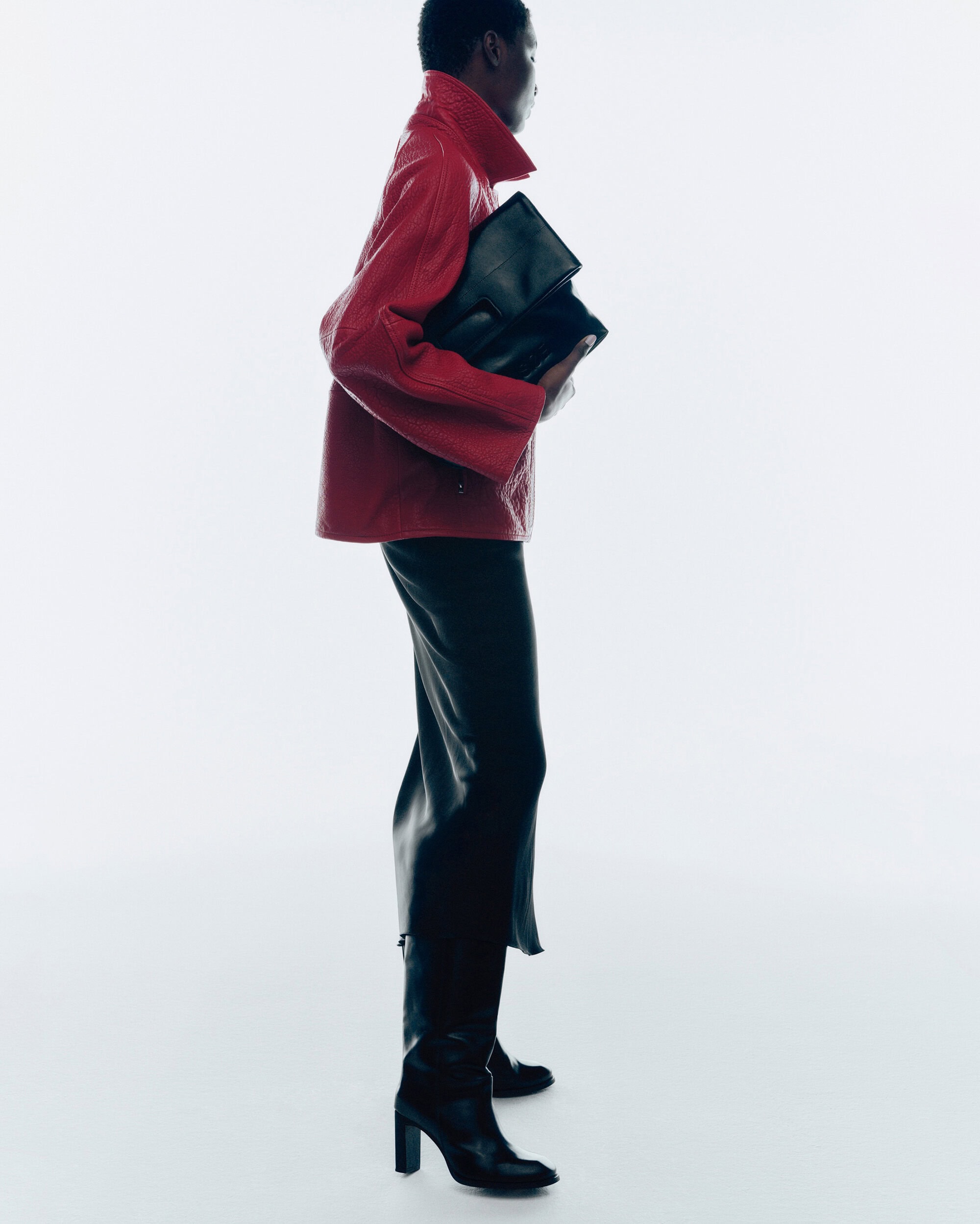 Model in red leather jacket and black skirt holding a black clutch against a white background