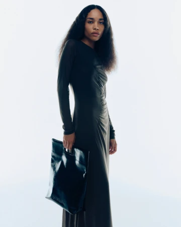 Model in dark long-sleeved dress holding a black leather tote bag against a plain white background for Samsoe Samsoe campaign