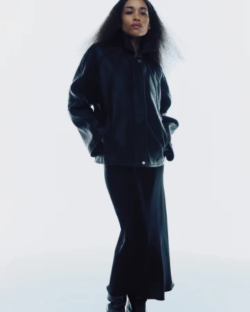 Model wearing a black leather jacket and long black skirt against a white studio background for Samsoe Samsoe.