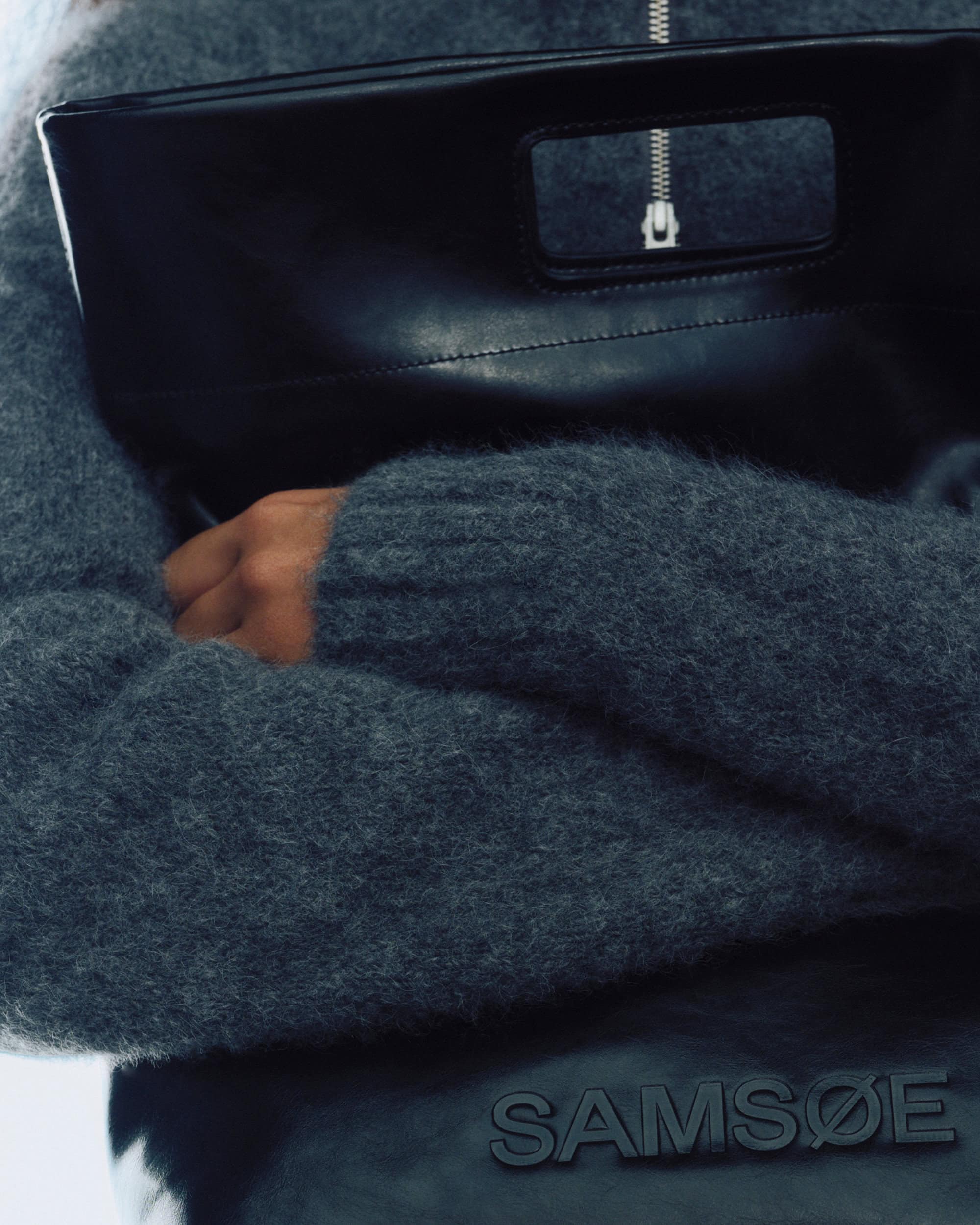 Close-up of a person wearing a textured grey knit sweater holding a smooth black leather handbag