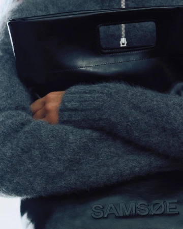 Close-up of a person wearing a textured grey knit sweater holding a smooth black leather handbag
