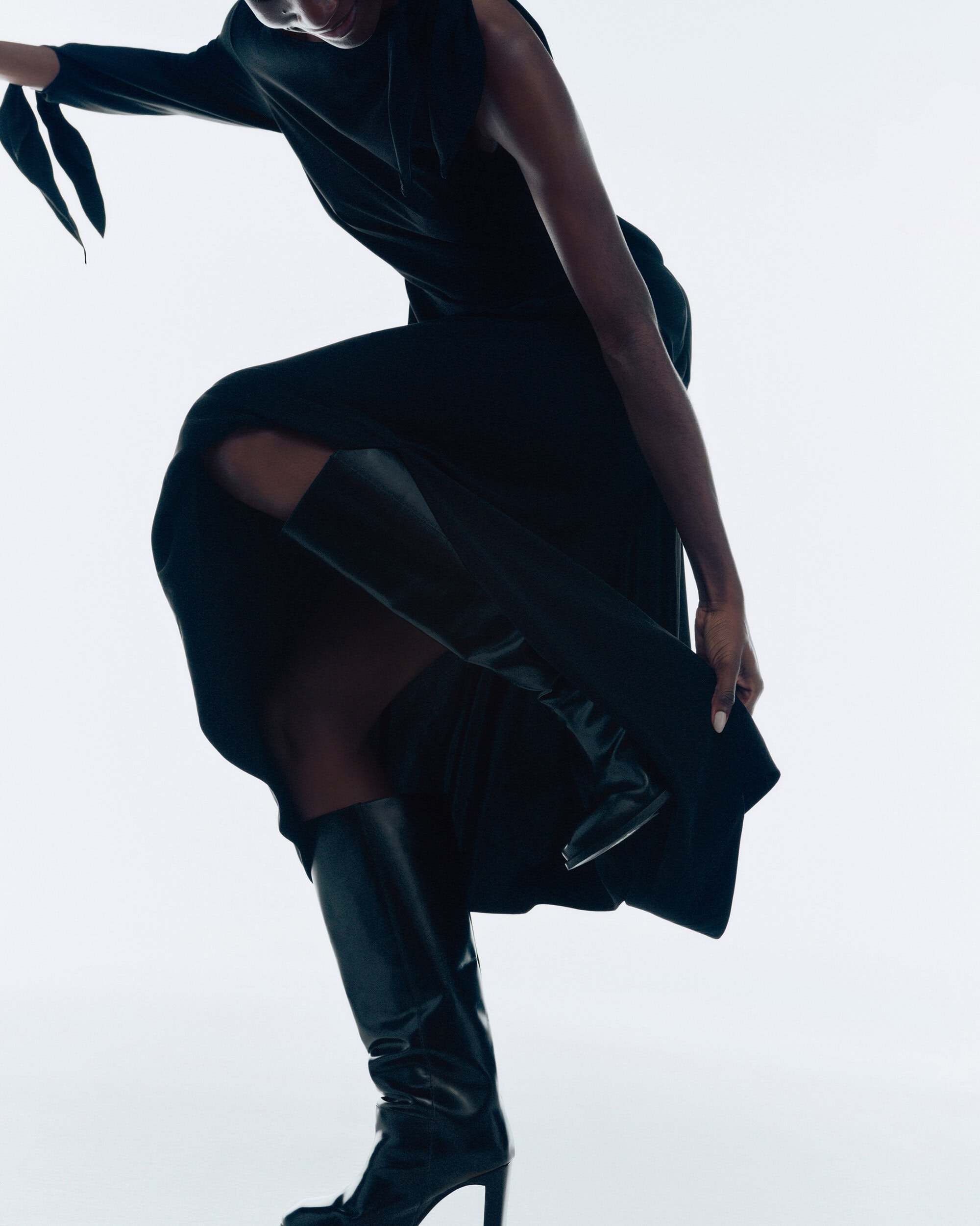 Model in a black dress and high leather boots lifting one leg against a bright white background