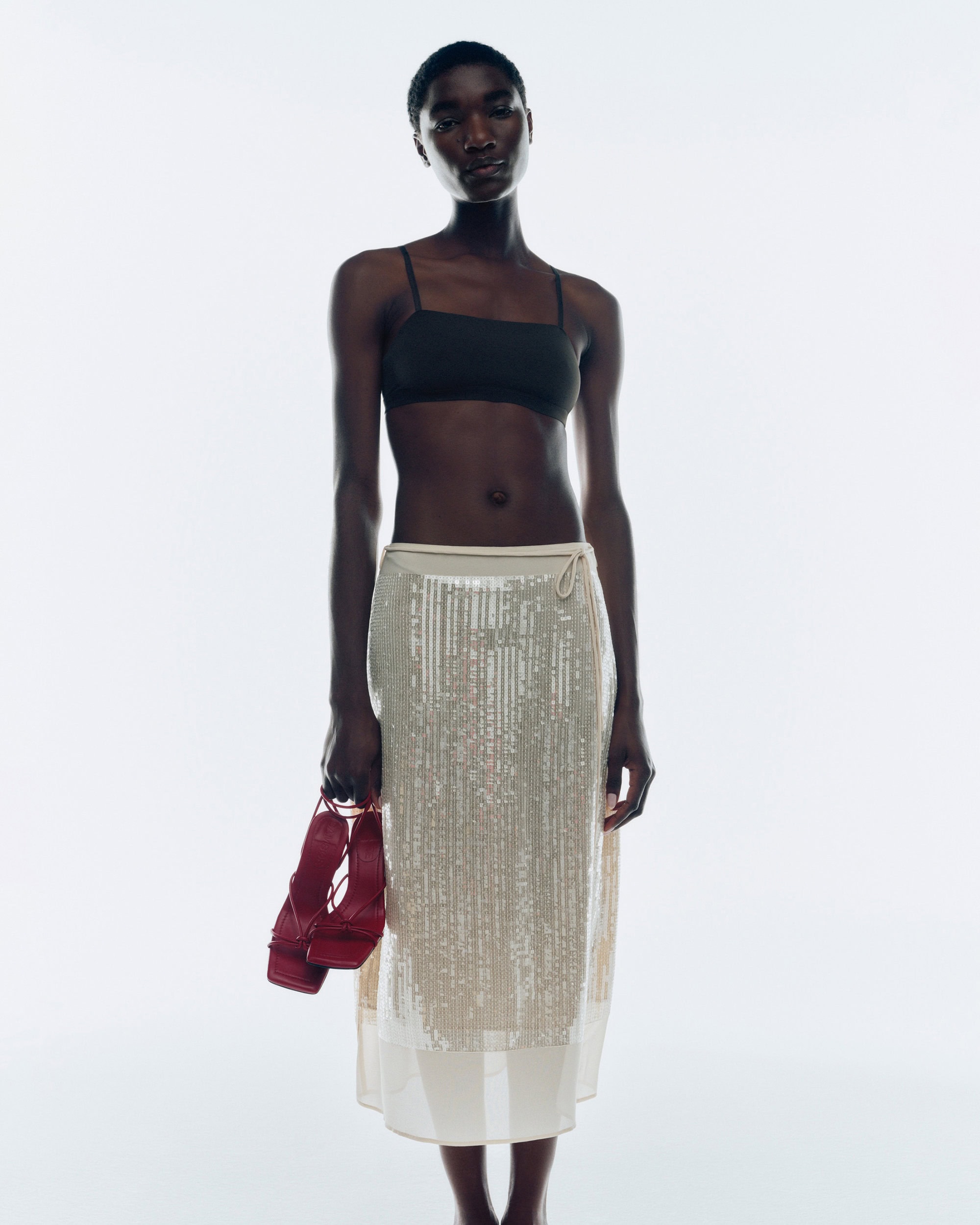 Model standing against a white backdrop wearing a black top and a semi-sheer skirt, holding red sandals