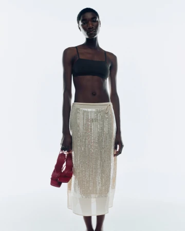 Model standing against a white backdrop wearing a black top and a semi-sheer skirt, holding red sandals