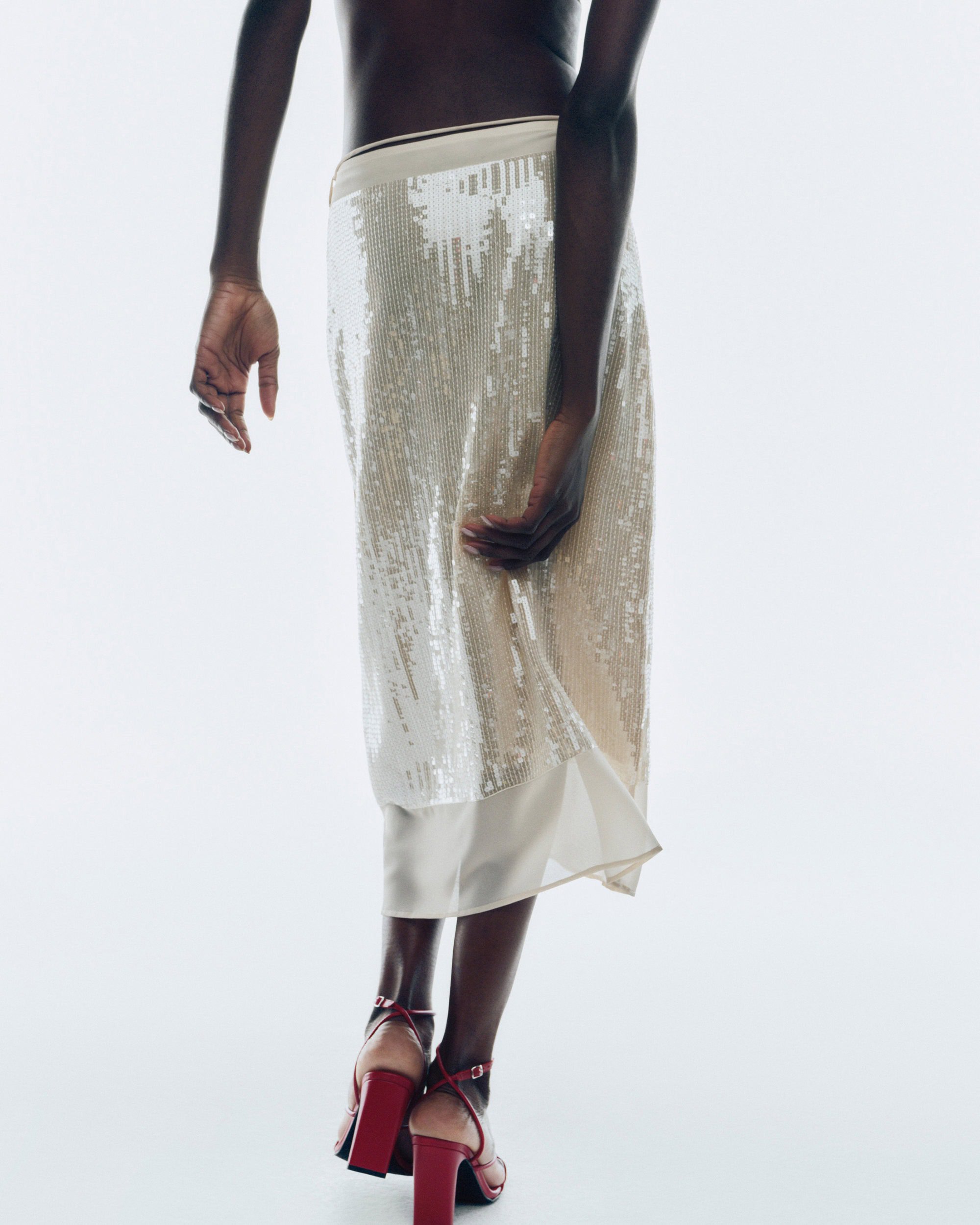 Model wearing a sequined skirt and red heels photographed from behind against a white background