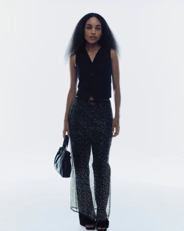 Model wearing a black vest and sequin trousers holding a black handbag against a white background