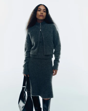Model wearing a dark grey knit zip-up cardigan and matching skirt holding a black leather handbag against a white background