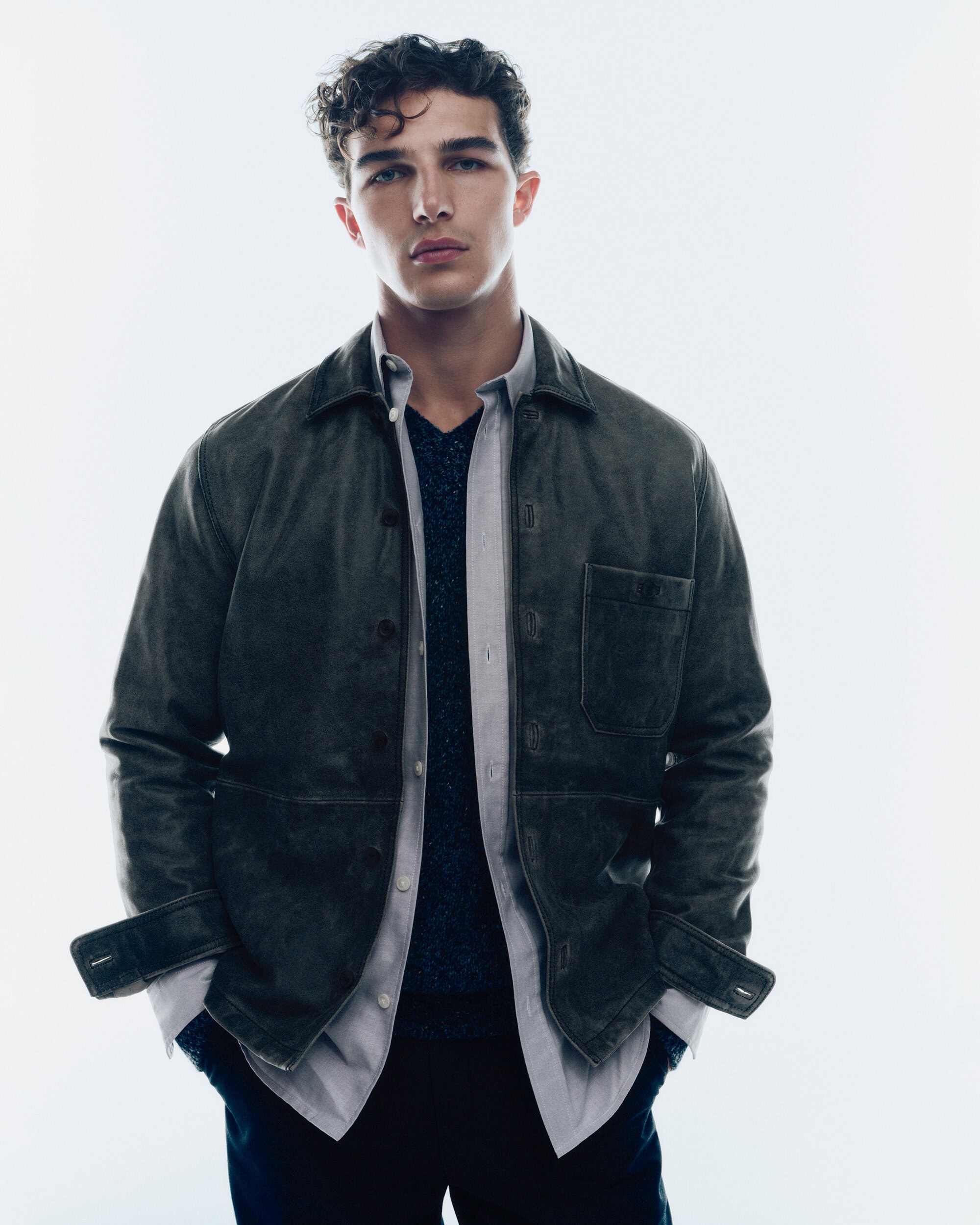 A man wearing a dark olive jacket and open grey shirt against a plain white background for Samsøe Samsøe pre-spring 2025 ad campaign.