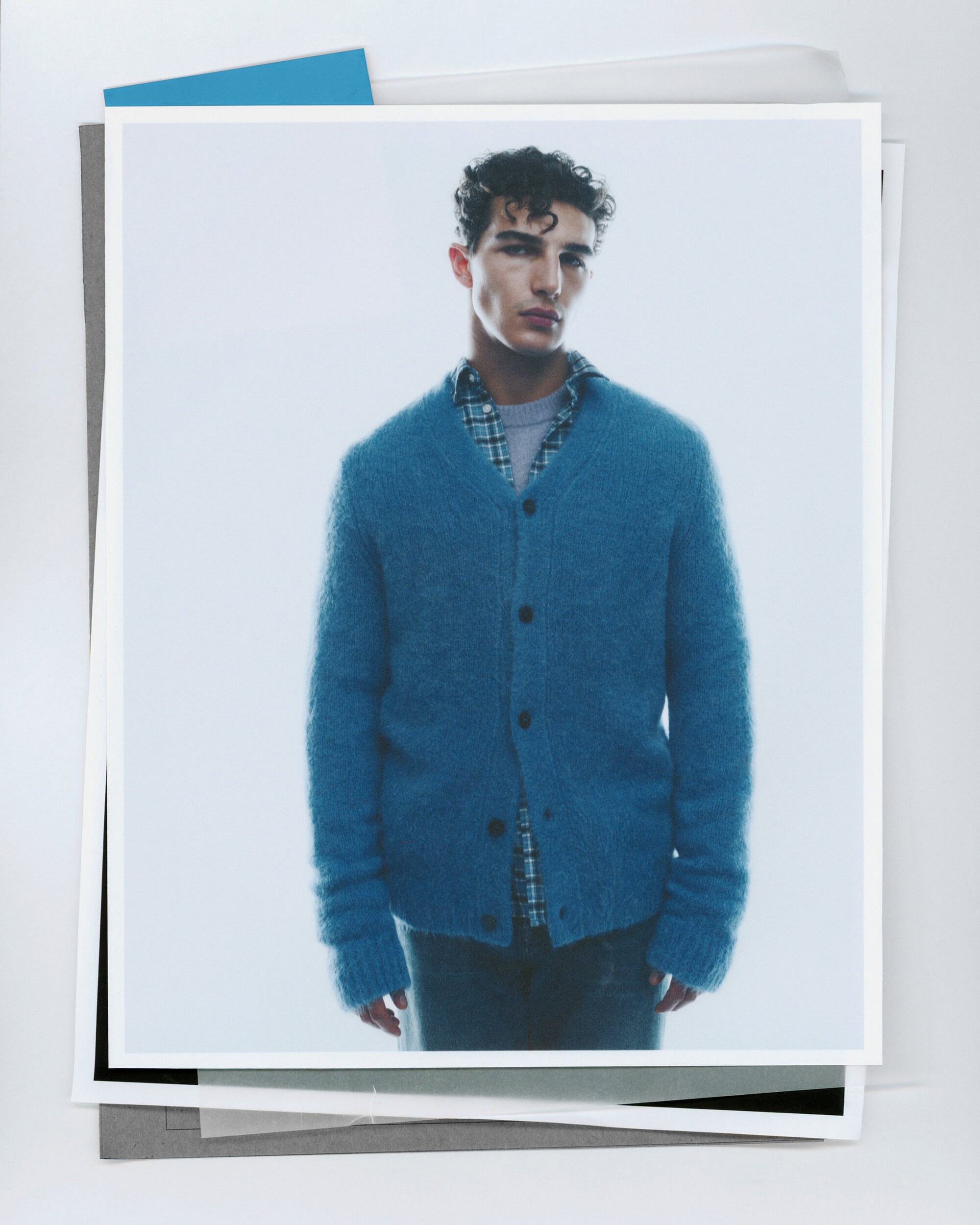 A young man wearing a blue knit cardigan over a plaid shirt against a minimalist white background for Samsoese Samsoe.