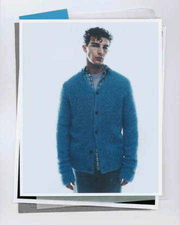 A young man wearing a blue knit cardigan over a plaid shirt against a minimalist white background for Samsoese Samsoe.