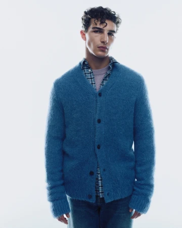 A man wearing a blue fuzzy cardigan over a plaid shirt and lavender sweater against a plain white background.
