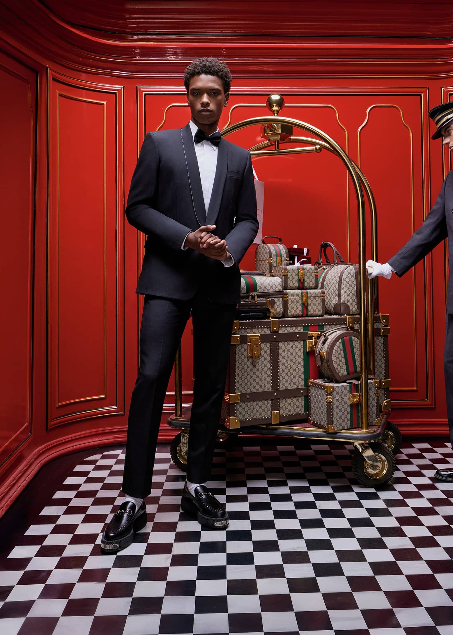 Man wearing a black tuxedo and bow tie standing in front of luggage on a gold cart inside a red room