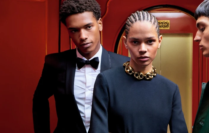 Group of three models in formal attire and heavy gold necklace against a vibrant red background for Gucci Holiday 2024 campaign