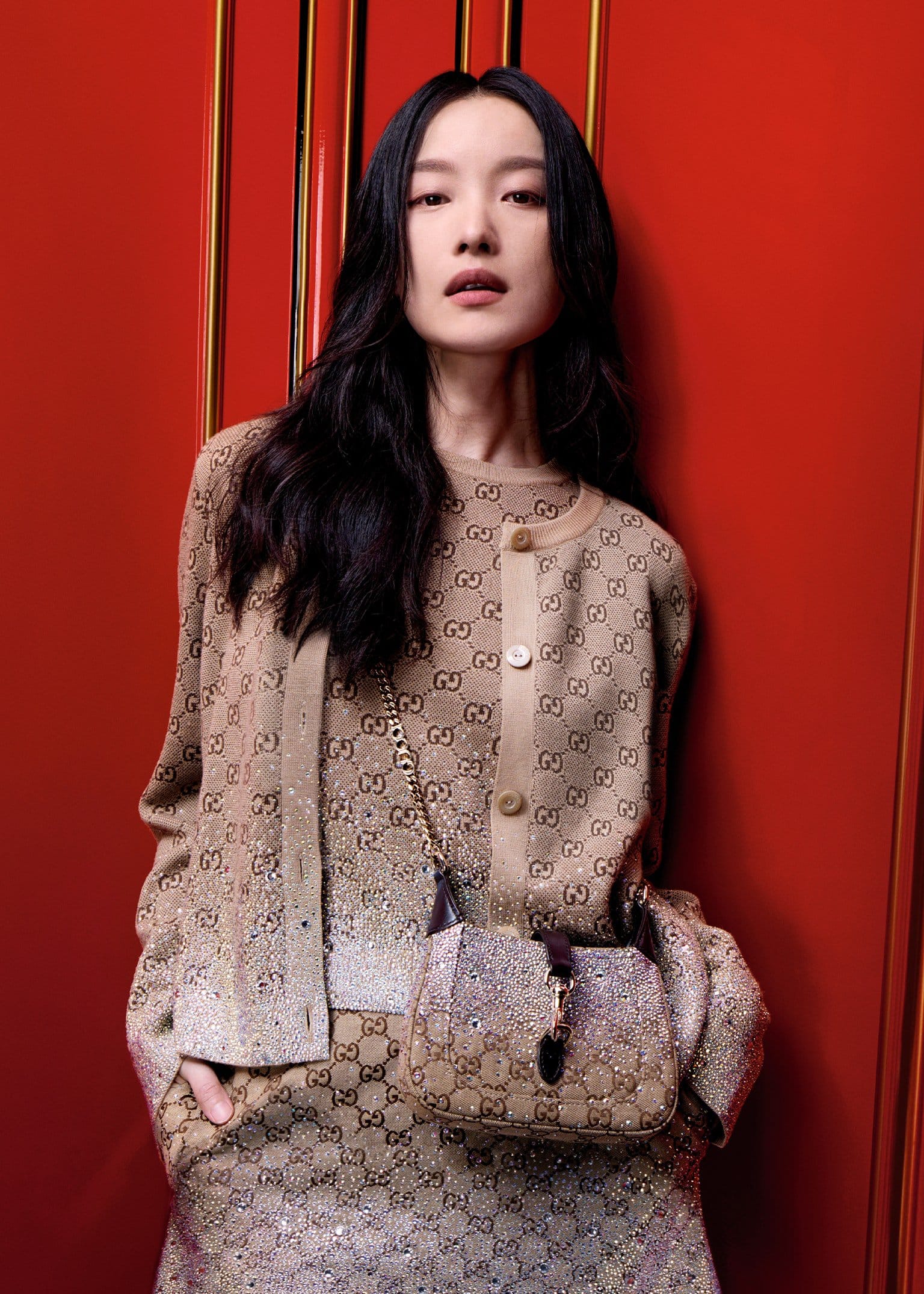 Woman in monogram cardigan and skirt leaning against red wall wearing a shoulder bag for Gucci Holiday campaign
