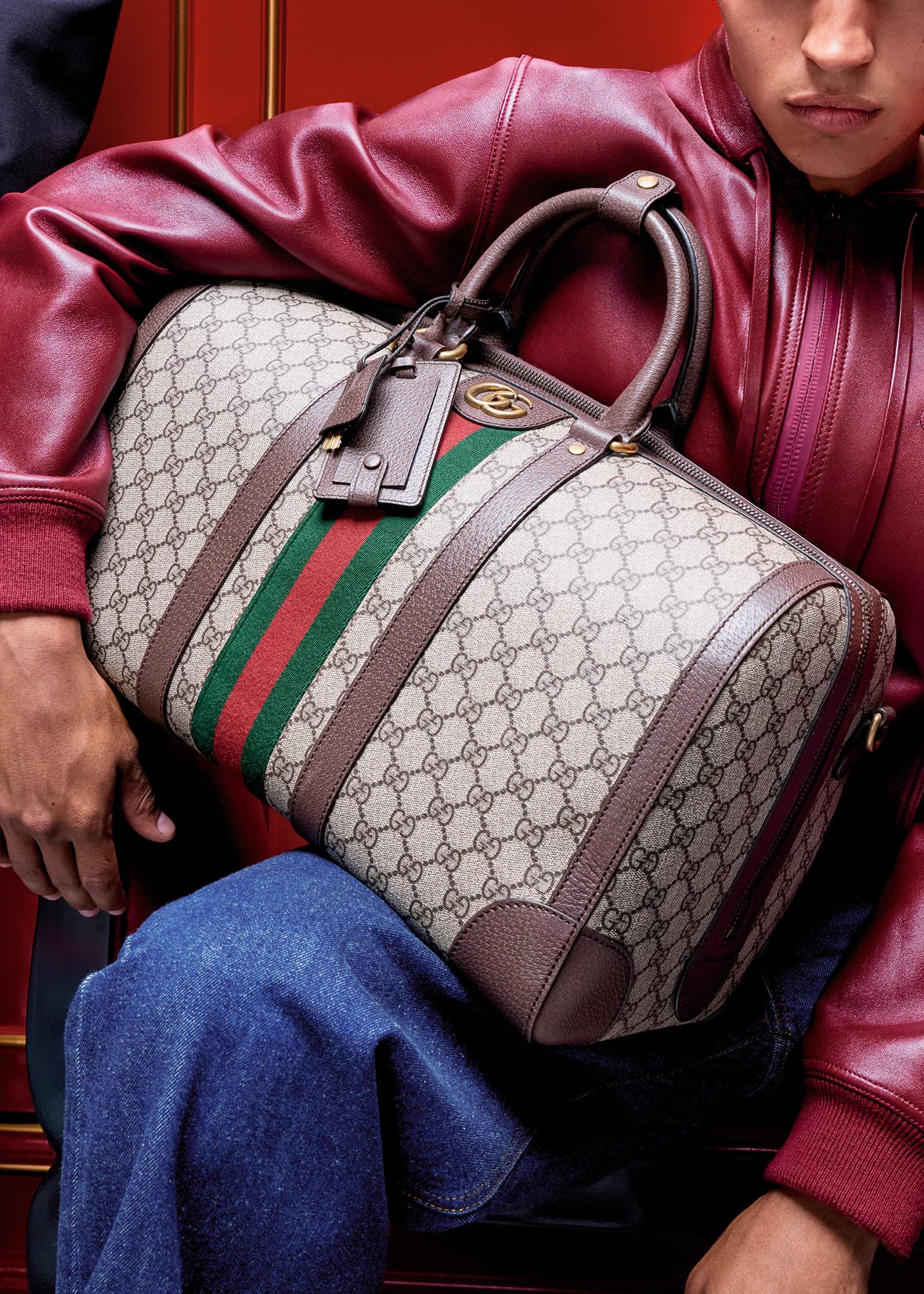 Close-up of a monogram duffle bag with green and red web stripe held by a person in a burgundy leather jacket and blue denim jeans.