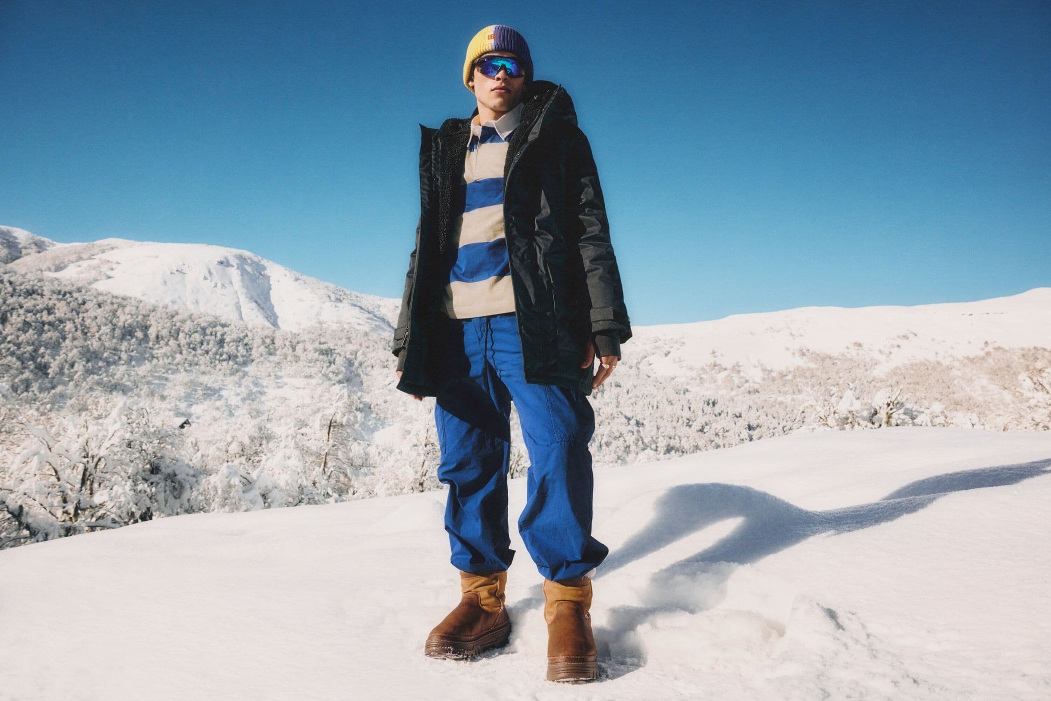 Model wearing a black puffer jacket, blue trousers, and sunglasses standing in a snowy landscape under a clear blue sky