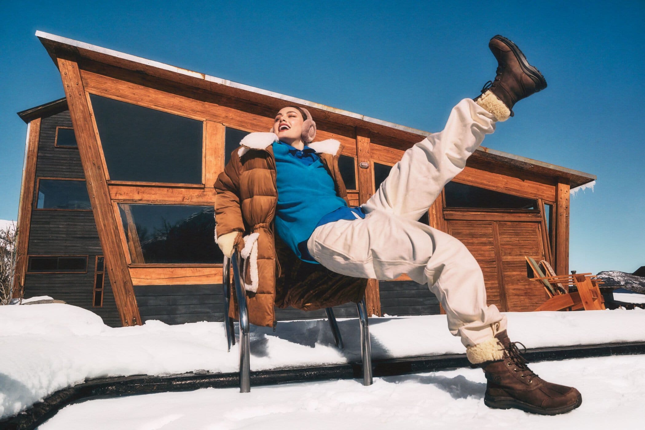 Woman in a brown winter coat and boots kicking her leg up outside a modern wooden cabin on snow.