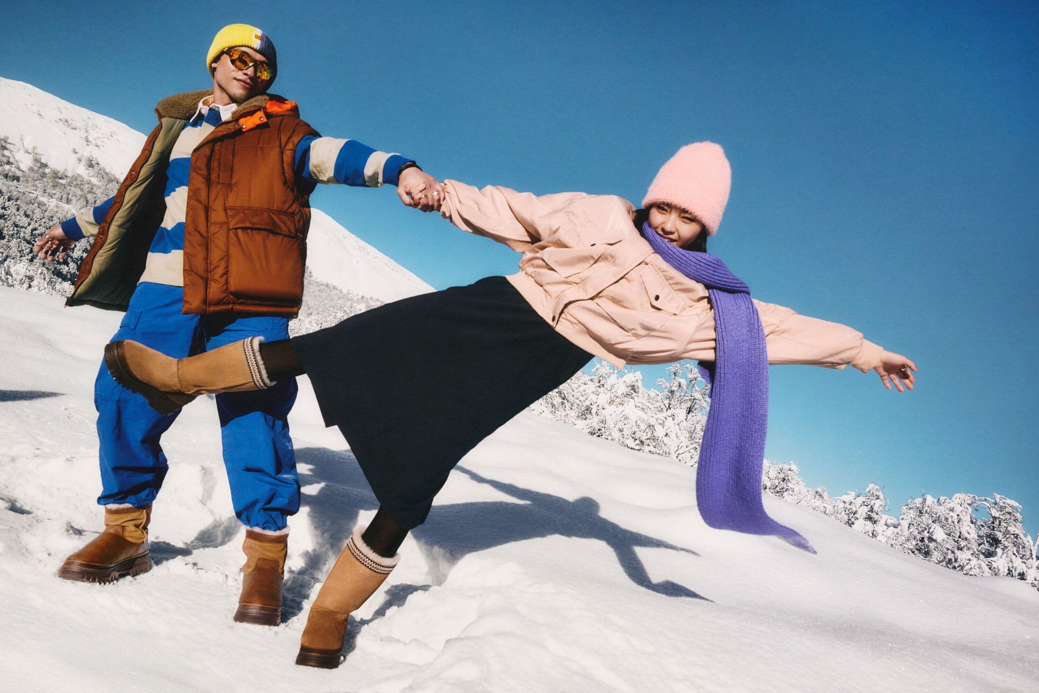 Two models posing outdoors in a snowy landscape wearing winter apparel and tan boots under a clear blue sky
