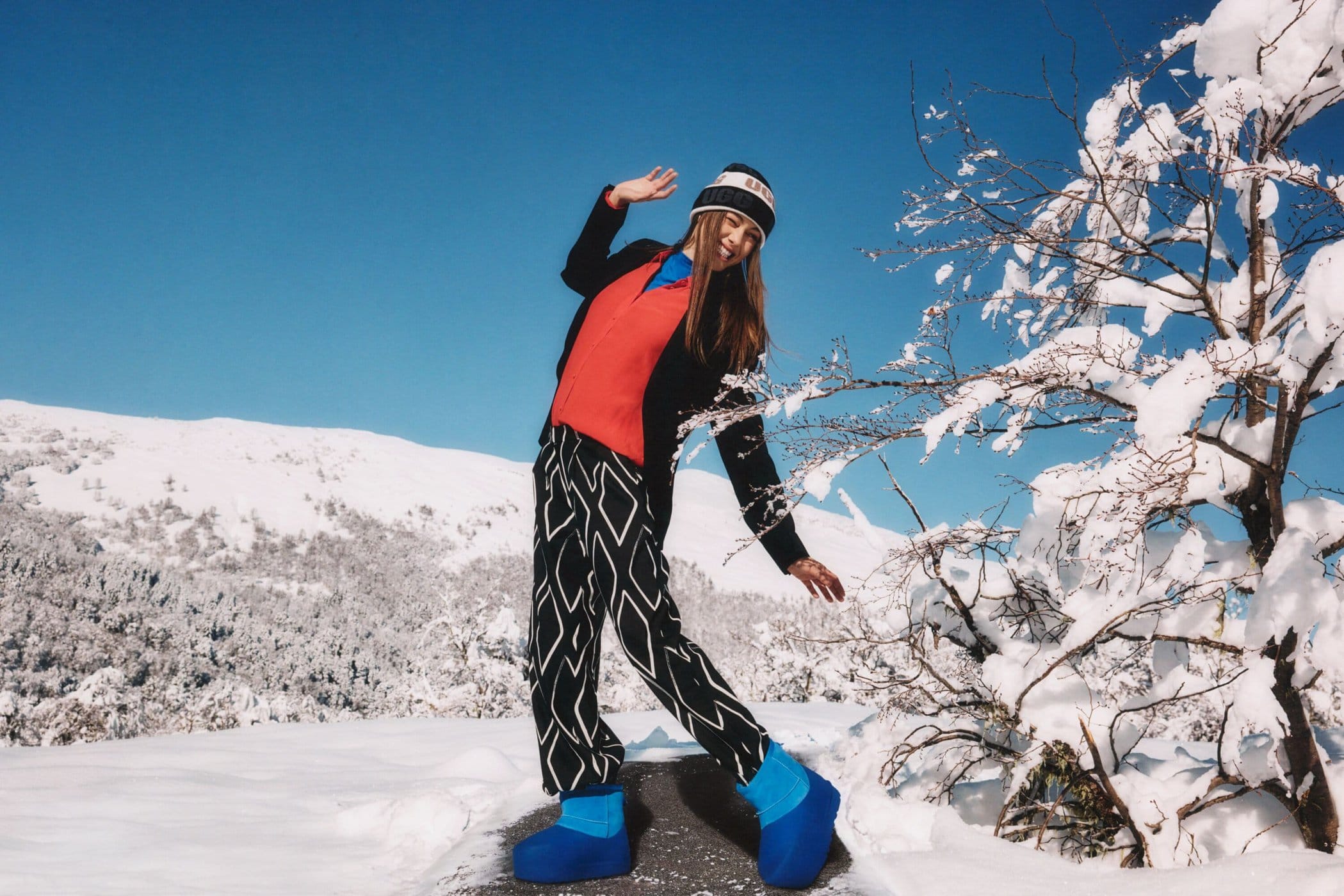 A smiling woman wearing a beanie and bright blue boots poses outdoors on a snowy mountain path under a clear blue sky.