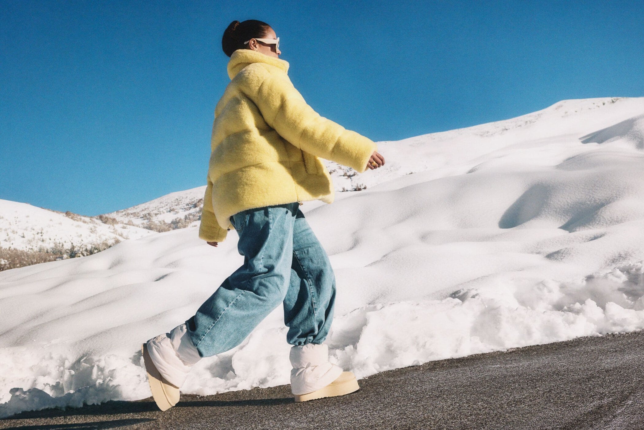 Woman in bright yellow fluffy jacket and blue jeans walking along a snowy mountain road under a clear blue sky