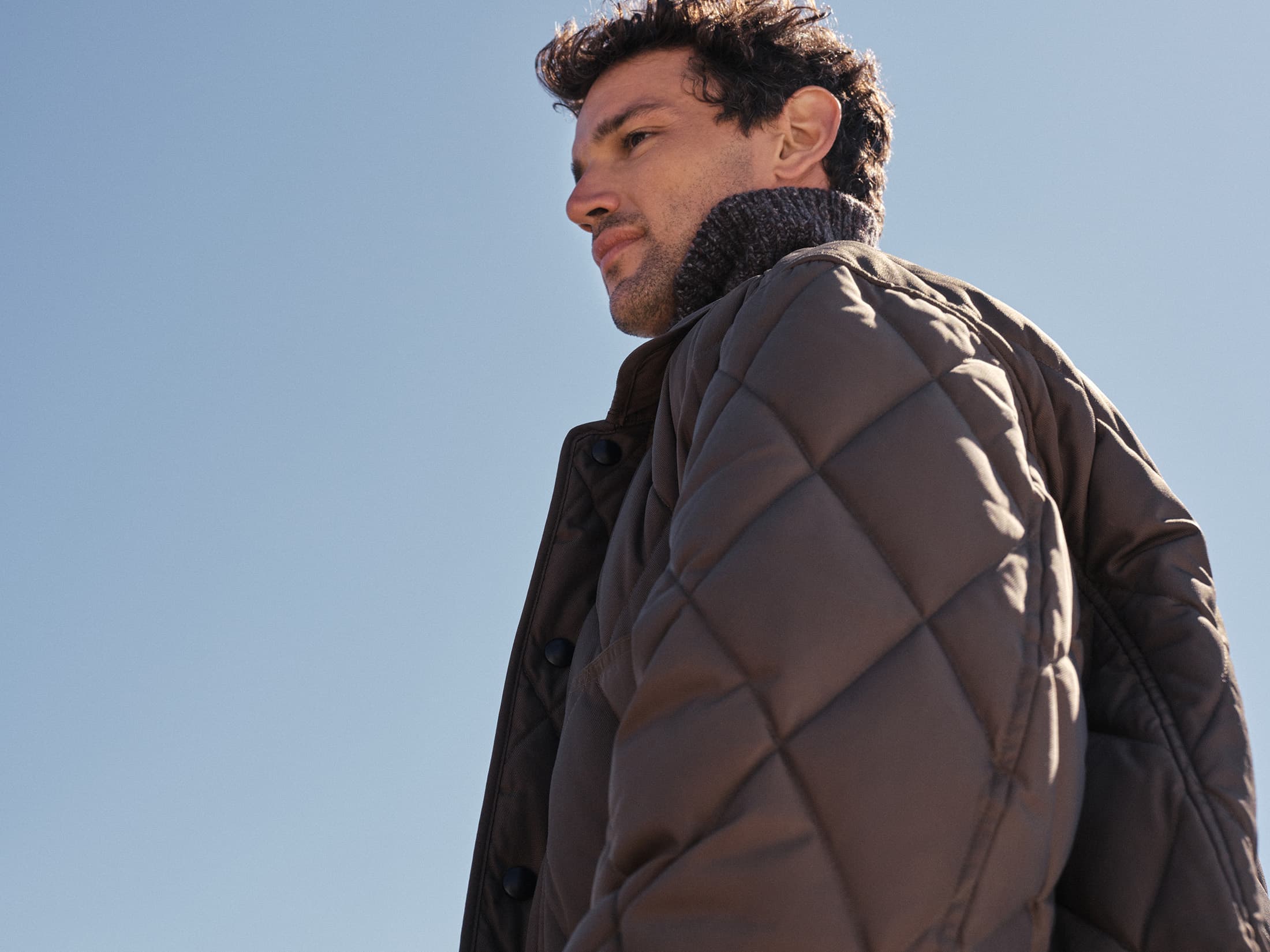 Man wearing a quilted brown jacket against a clear blue sky background