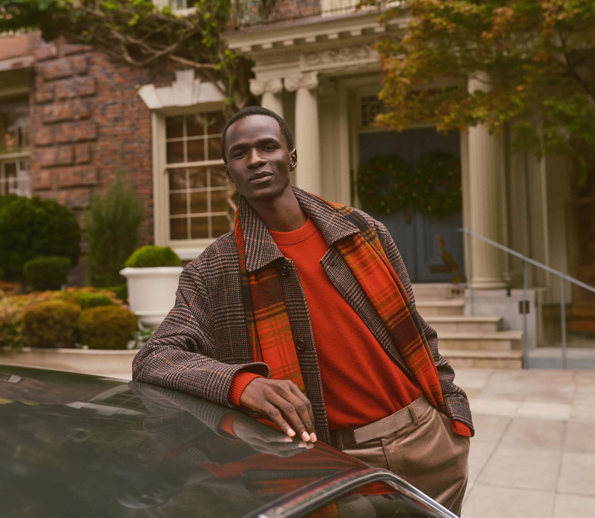 Black man wearing a plaid coat and red sweater leaning against a car in front of a residential building