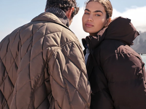 Close-up of a woman in a dark brown hooded jacket looking back, with a man wearing a quilted brown jacket turned away in the foreground.