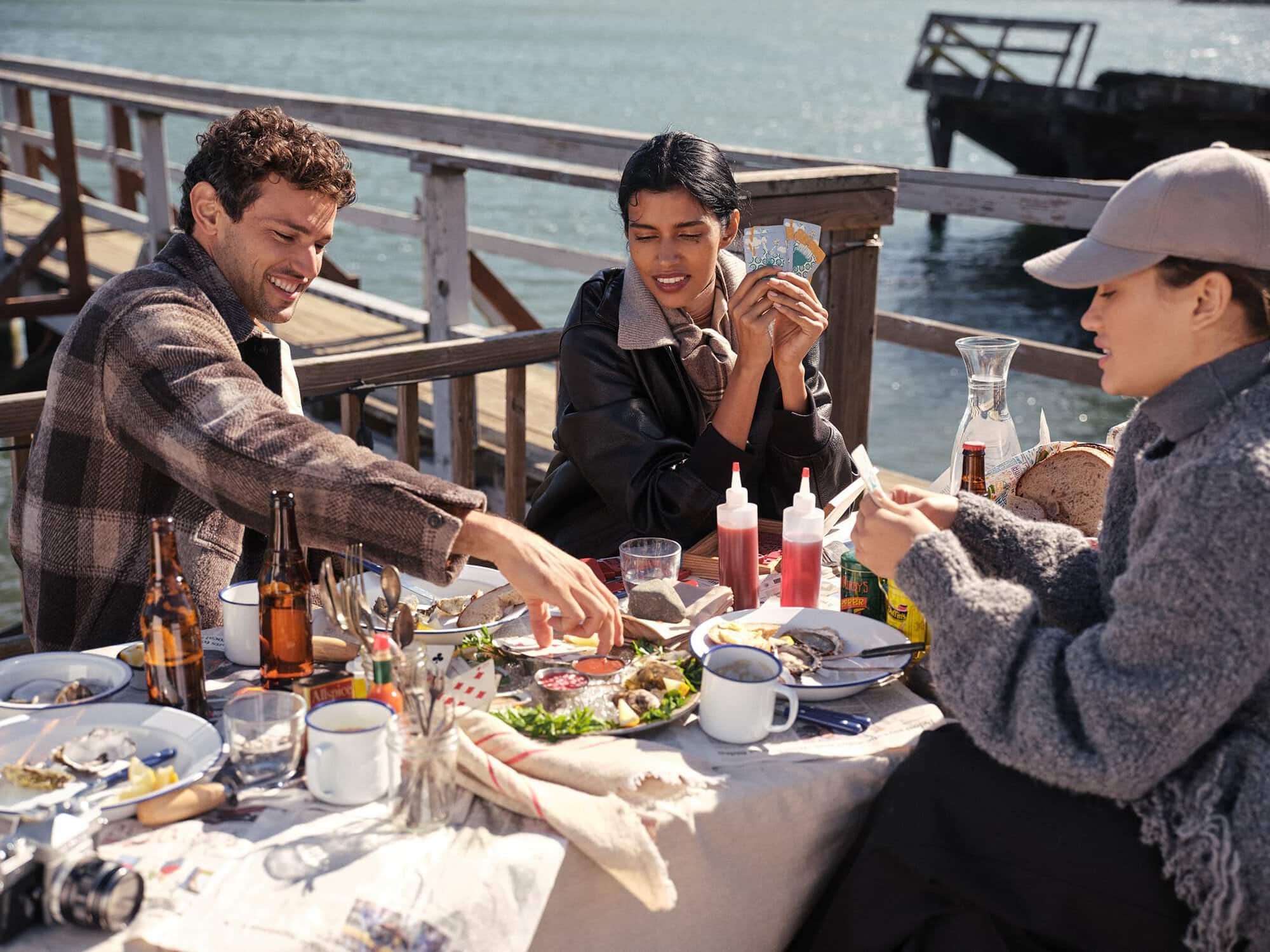 Three people sitting around a wooden outdoor table by the water, sharing a meal and playing cards.