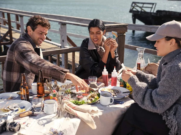 Three people sitting around a wooden outdoor table by the water, sharing a meal and playing cards.