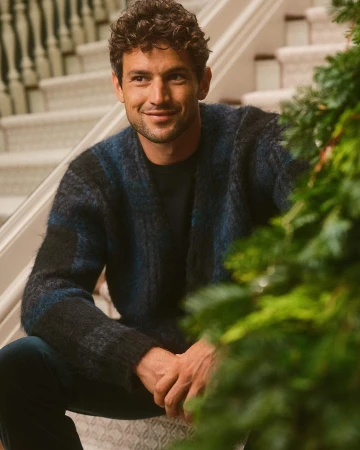 Man wearing a textured blue and black cardigan seated on indoor steps next to evergreen branches for Banana Republic.