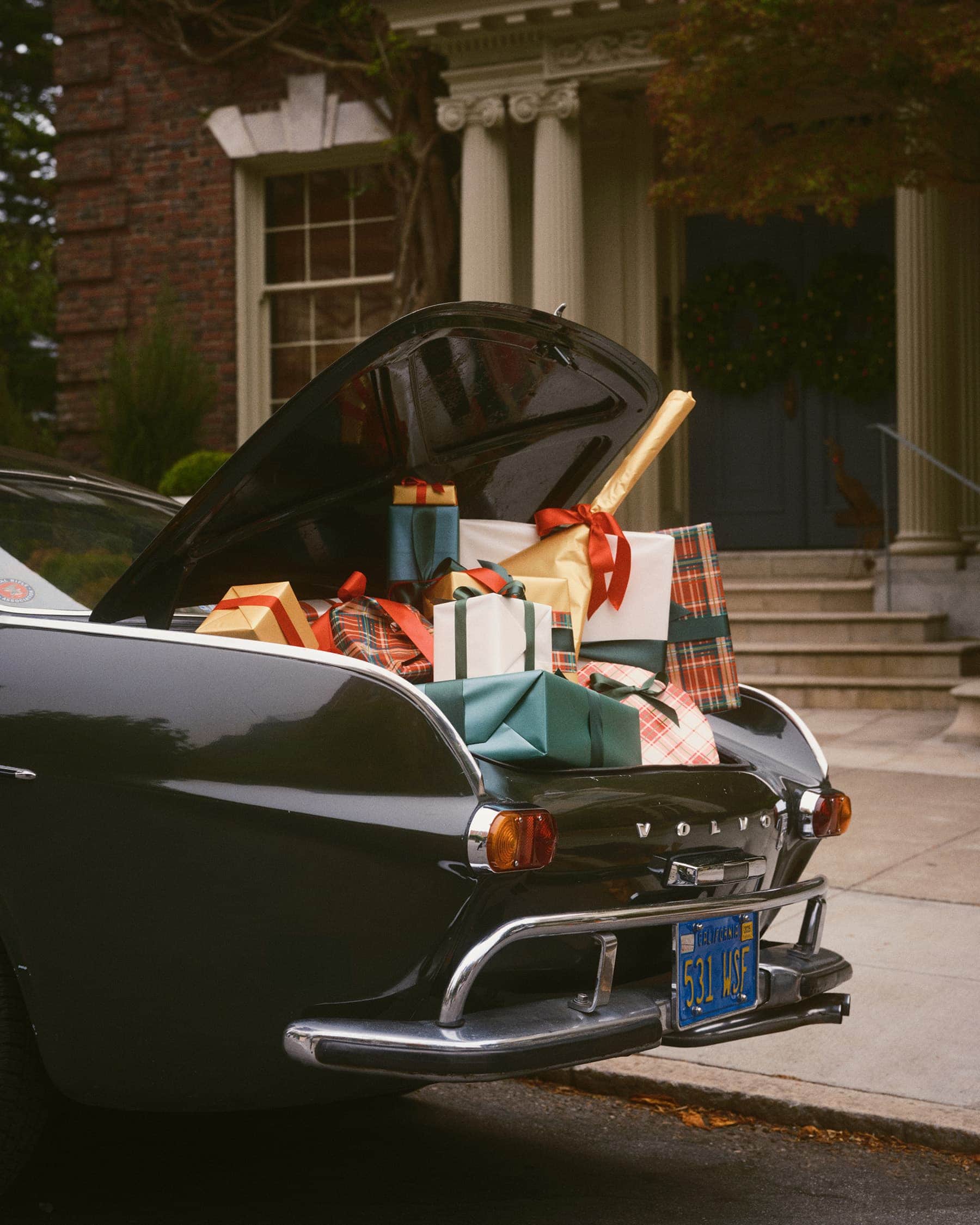 Trunk of a dark vintage car filled with wrapped holiday presents in front of a grand brick building.