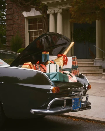 Trunk of a dark vintage car filled with wrapped holiday presents in front of a grand brick building.