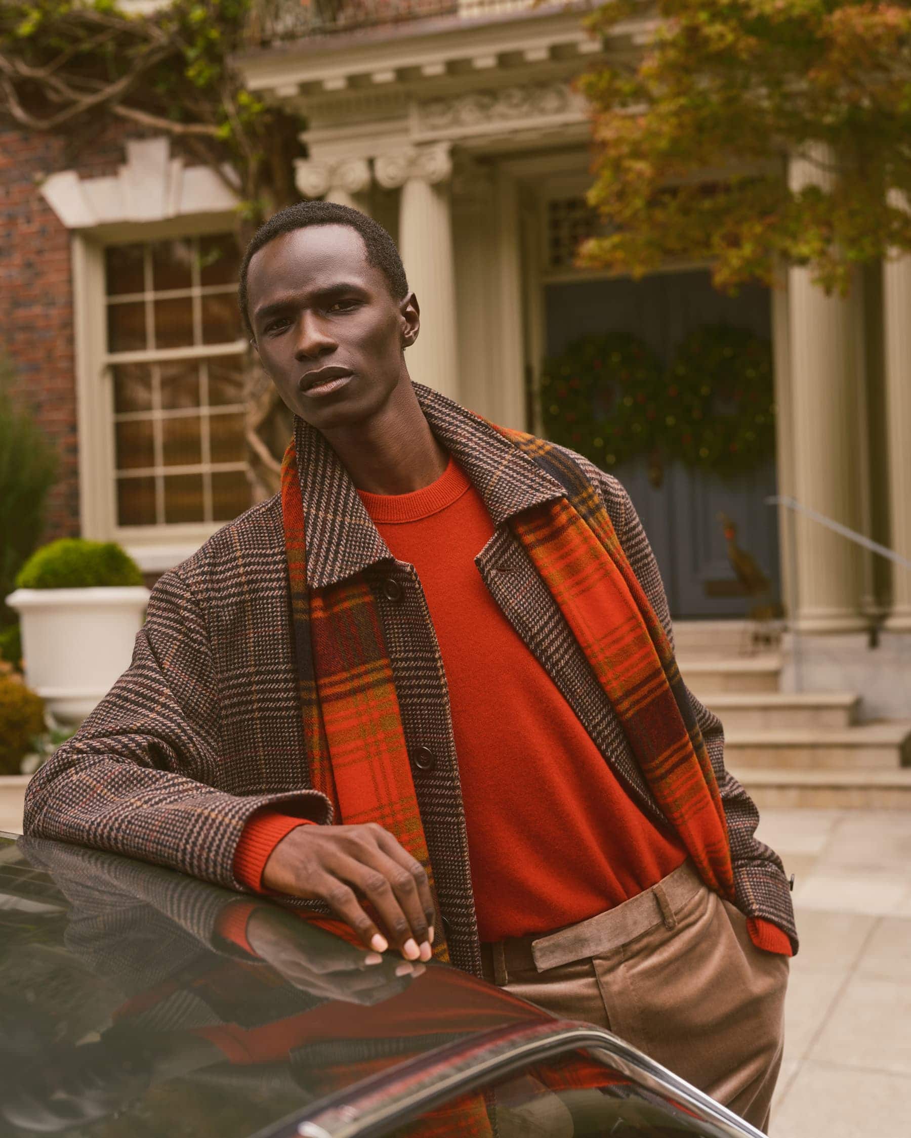 Black man wearing plaid coat, red sweater, and orange scarf leaning on car outside building