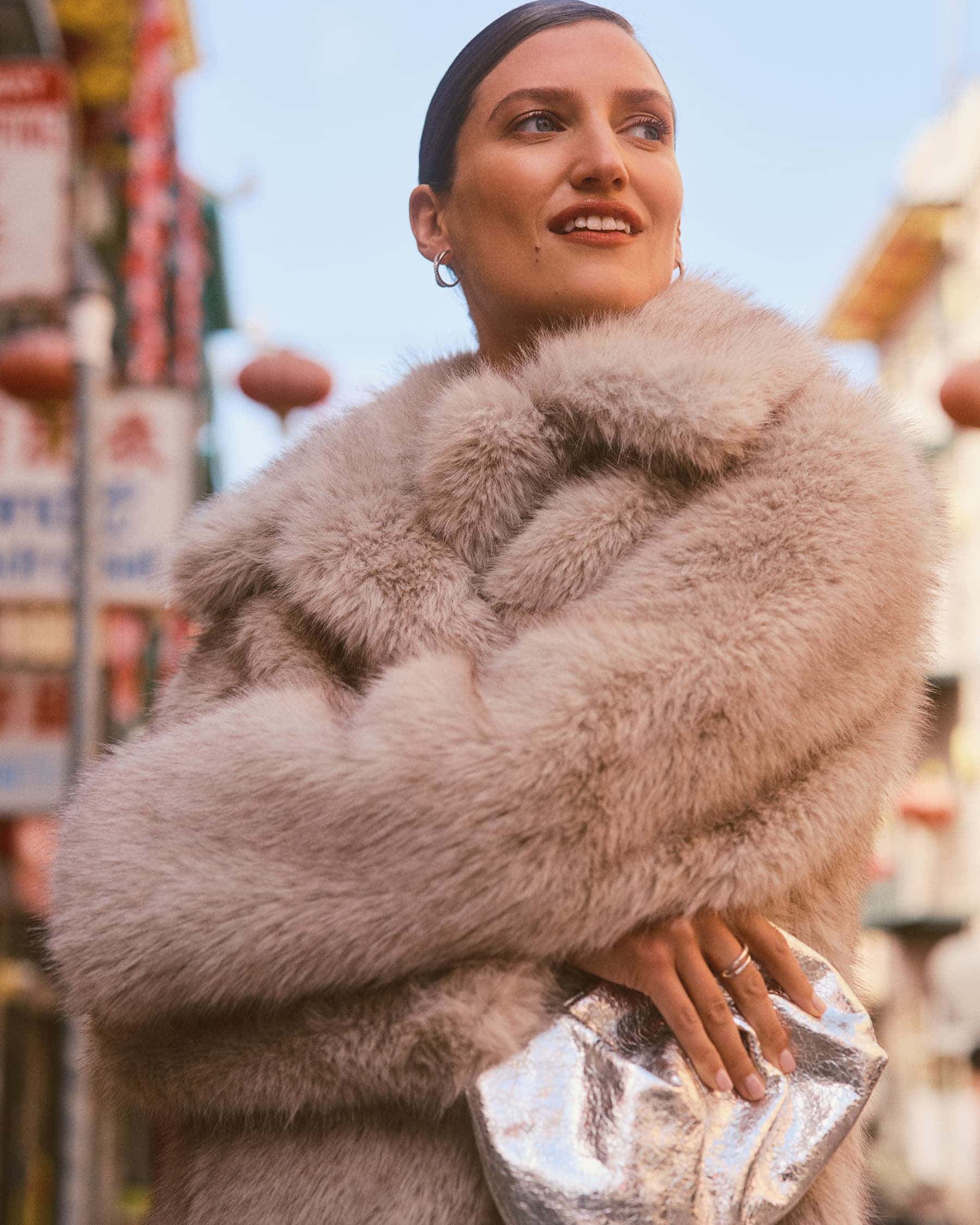 Woman wearing a beige faux fur coat and holding a silver metallic clutch outdoors on a city street