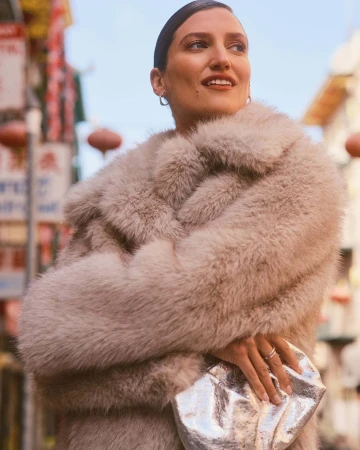 Woman wearing a beige faux fur coat and holding a silver metallic clutch outdoors on a city street