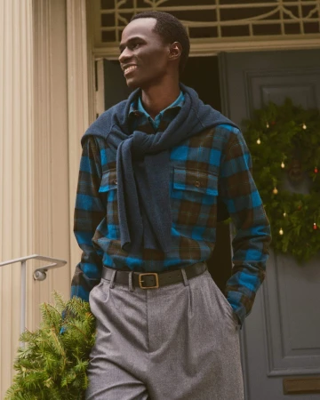 Man wearing a blue plaid shirt with a sweater tied over his shoulders and grey trousers outside a doorway.