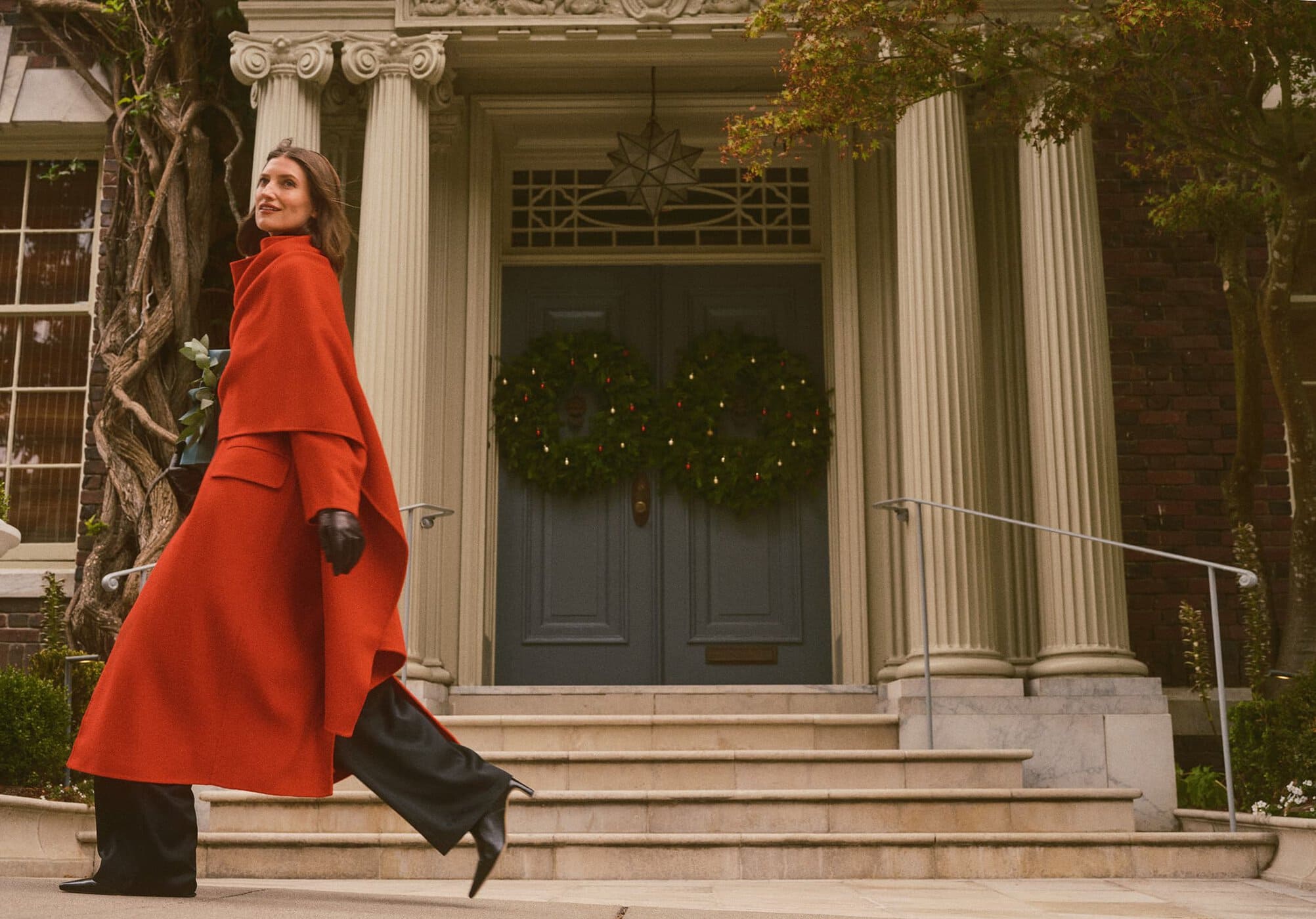 Woman in a long red coat and black leather gloves walking up steps outside a building with classical columns.