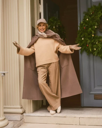 Elderly woman standing in doorway wearing beige sweater, trousers, and draped coat with holiday wreath