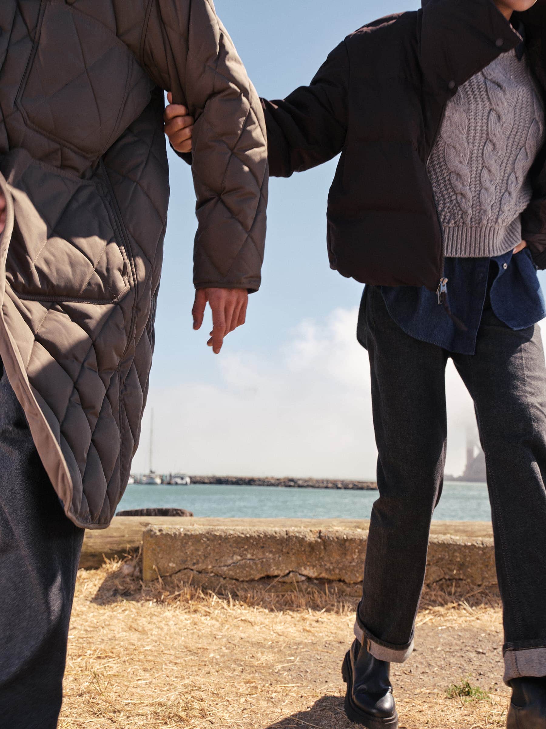 Two people wearing winter coats and trousers walking outdoors near a body of water during daytime.