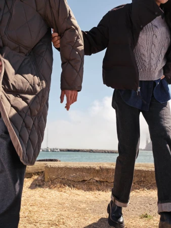 Two people wearing winter coats and trousers walking outdoors near a body of water during daytime.