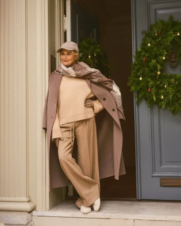 Lauren Hutton standing in an entryway wearing a neutral wool coat, sweater, trousers, and cap near a holiday wreath