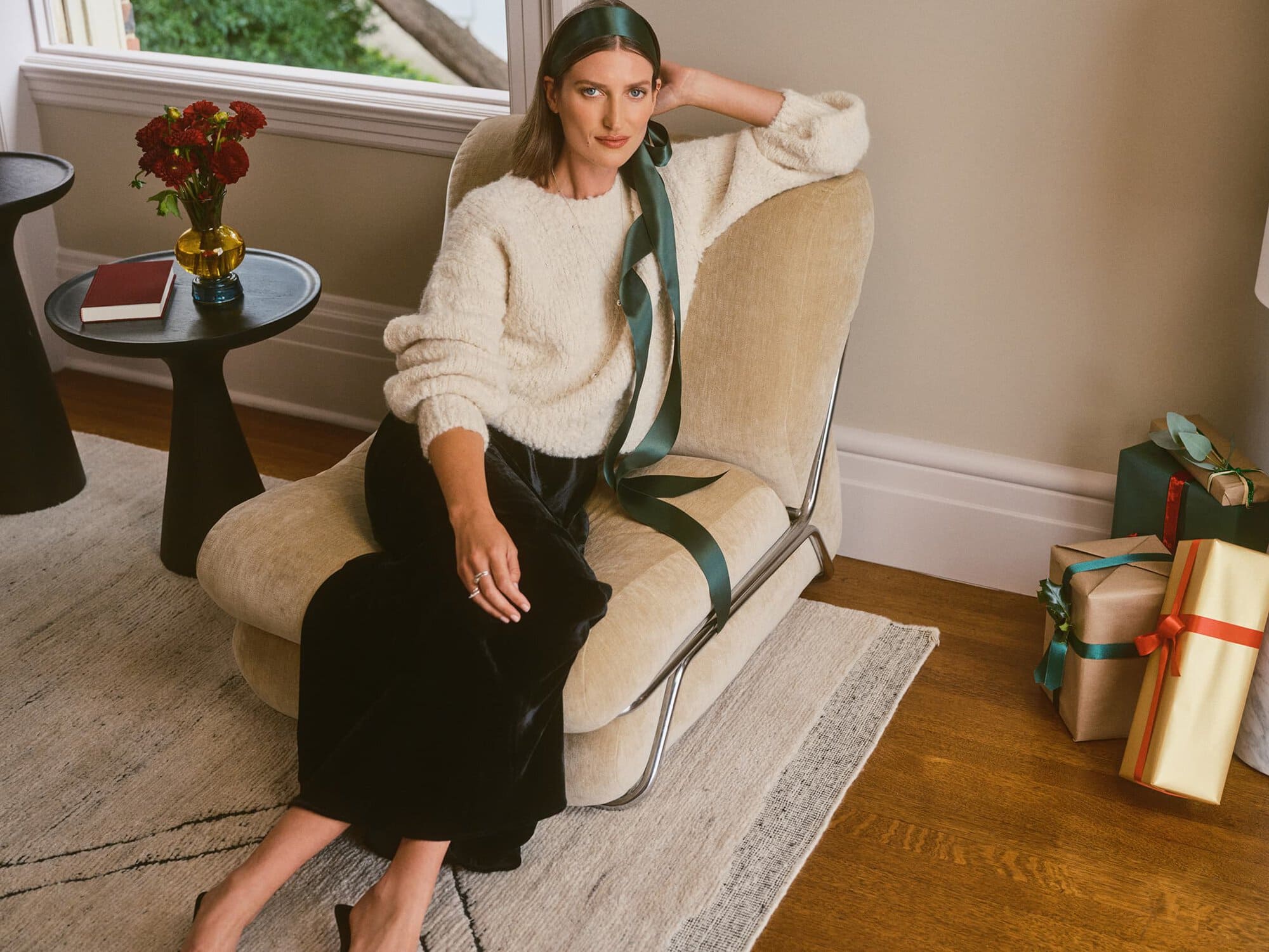 A woman wearing a cream sweater and black velvet skirt is seated on a beige armchair next to wrapped holiday gifts.