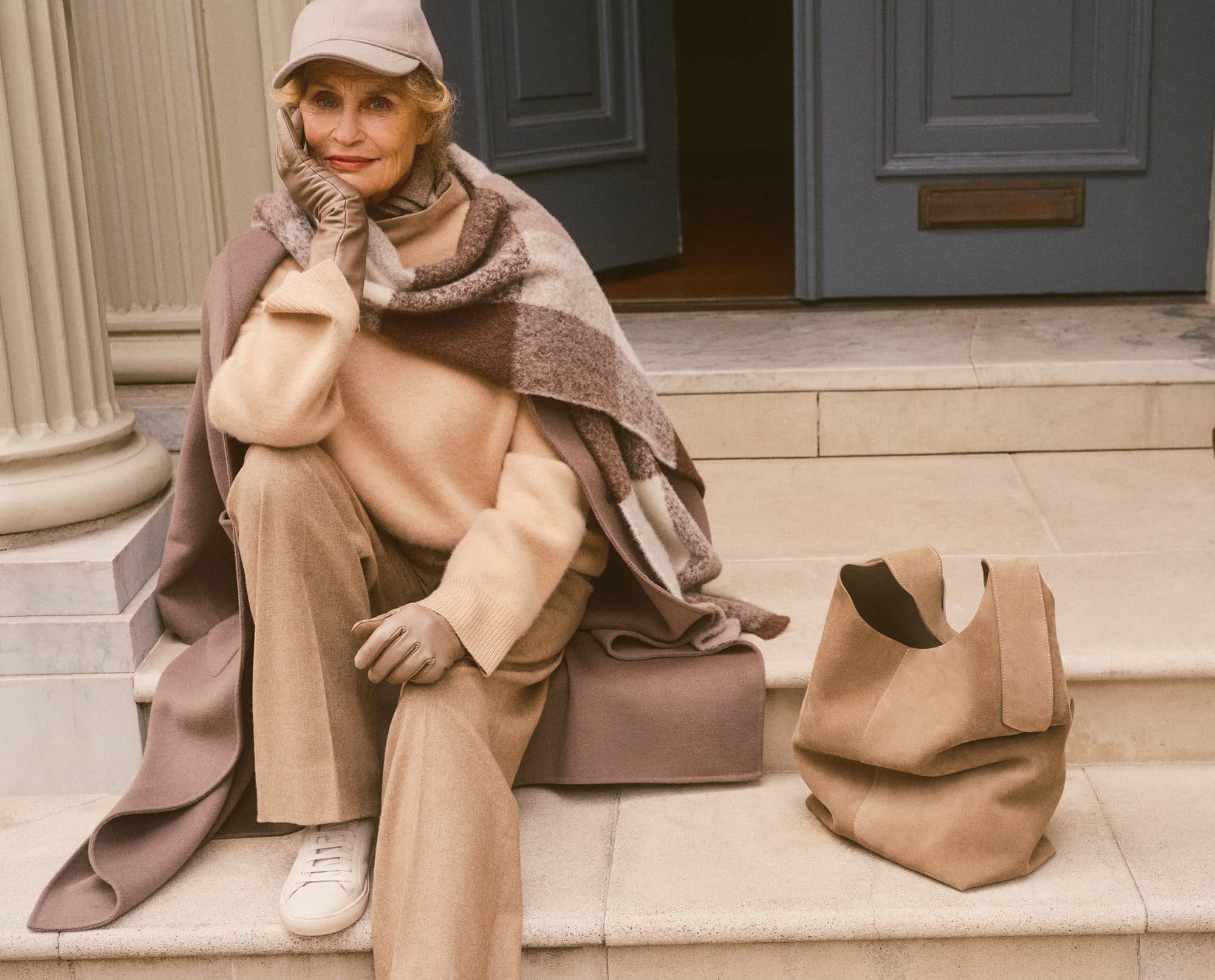 Elderly woman wearing a cap, sweater, trousers, and scarf seated on stone steps next to a leather handbag