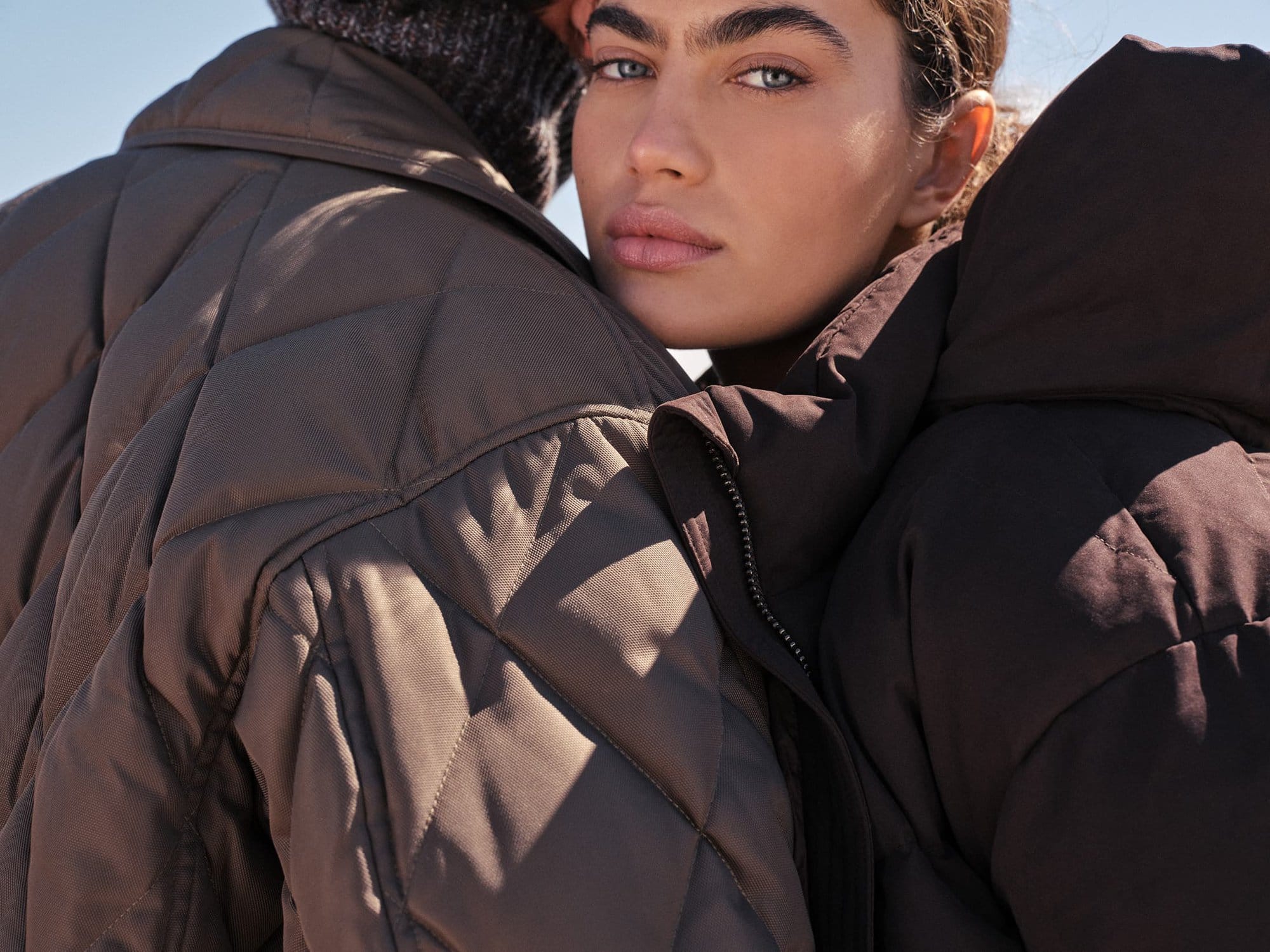 Close-up portrait of two models wearing quilted puffer coats against a bright clear sky background.