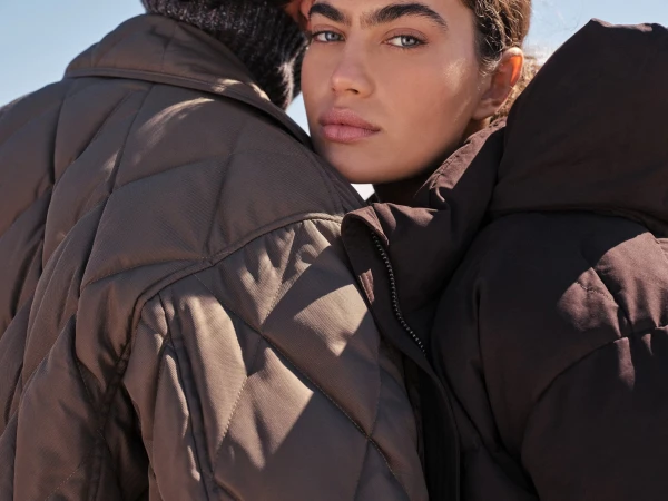 Close-up portrait of two models wearing quilted puffer coats against a bright clear sky background.