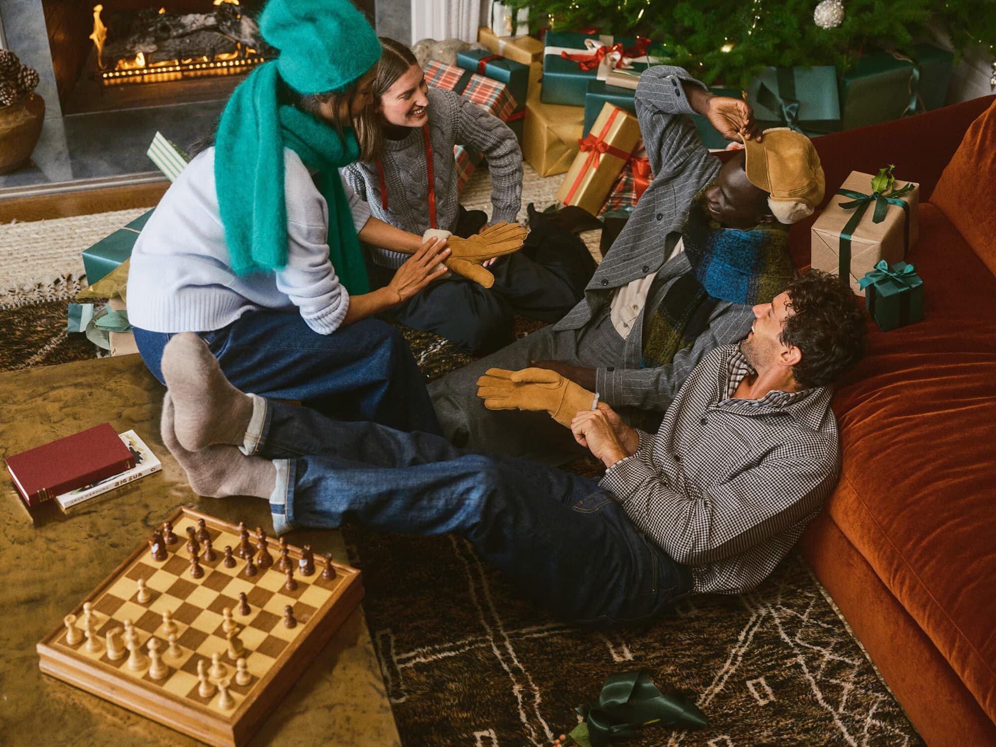 Group of four friends gathered around a living room opening Christmas presents, wearing winter knitwear and holding gloves.