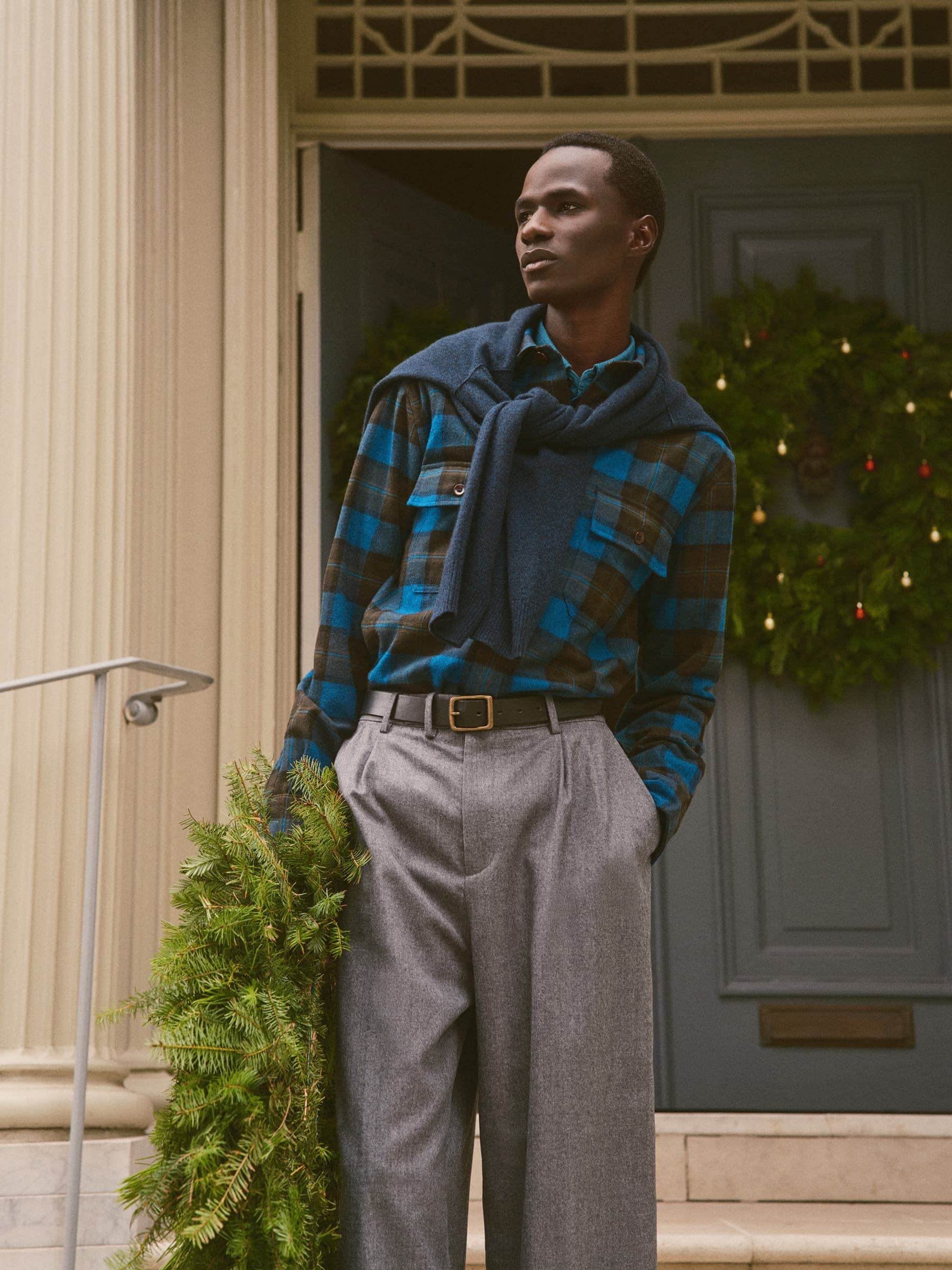 Black man standing on steps holding evergreen foliage in front of a house door with a Christmas wreath