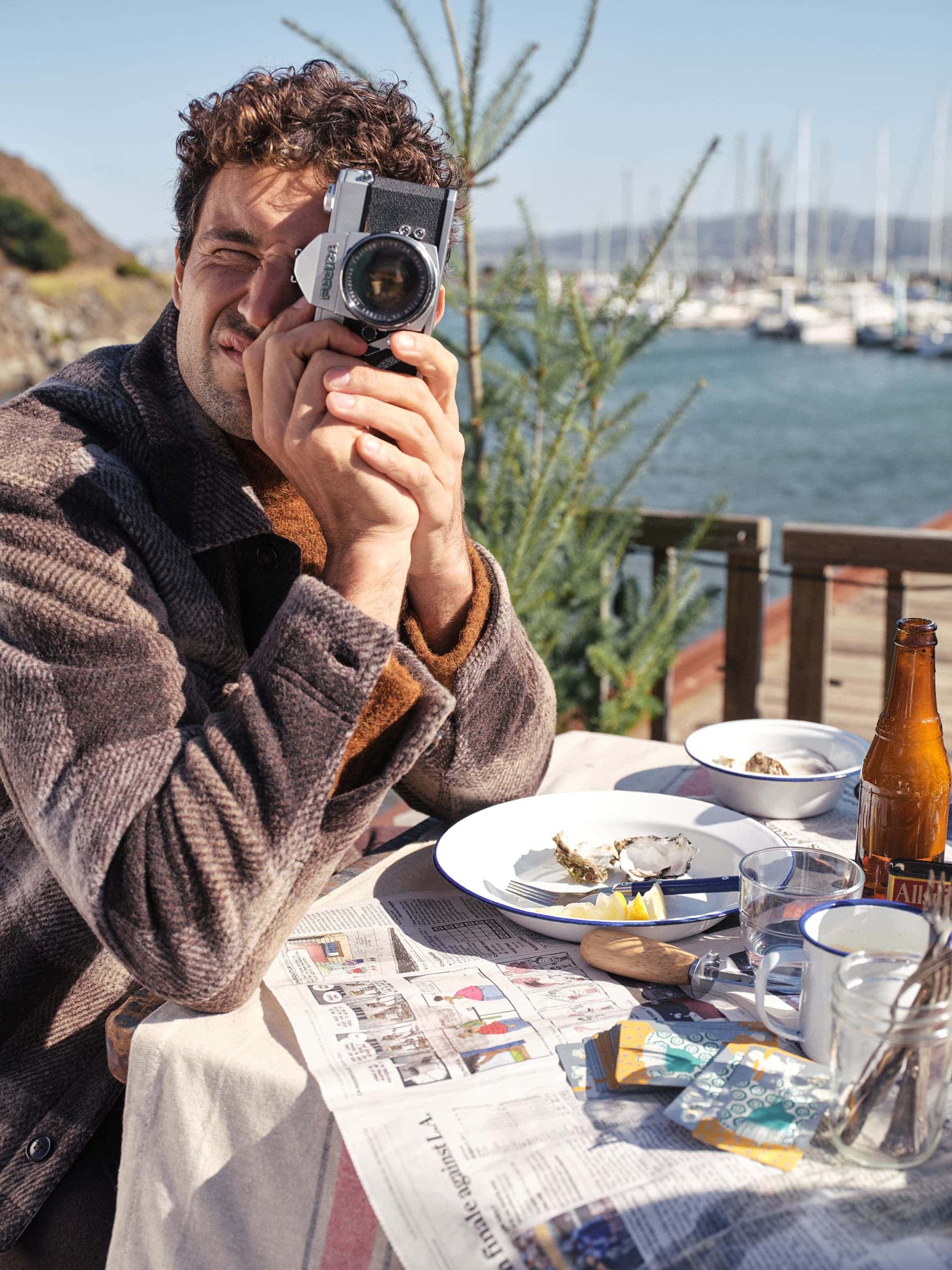 Man wearing a brown textured coat holding a vintage camera to his eye at an outdoor table by a marina.